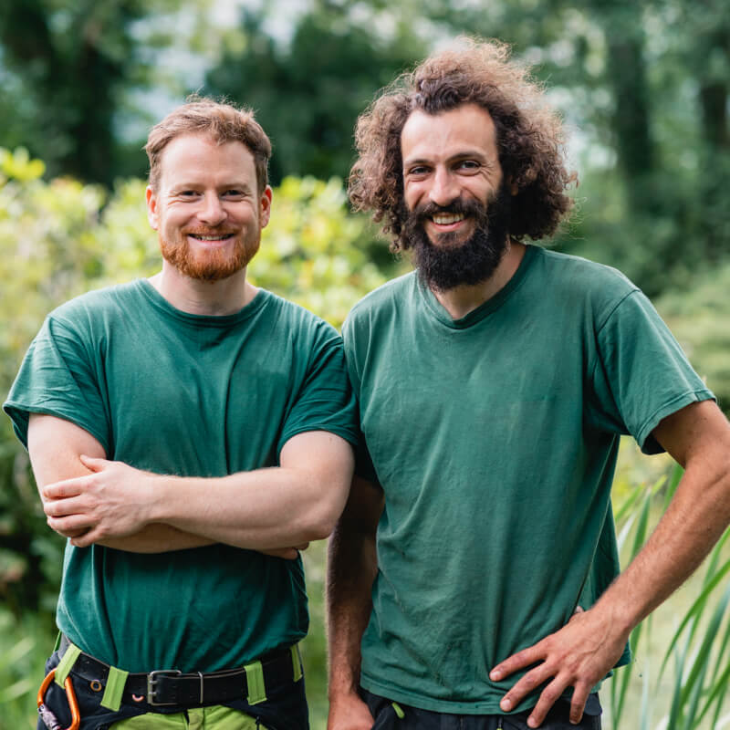 Zwei Landschaftsgärtner schauen lächelnd im Grünen stehend in die Kamera